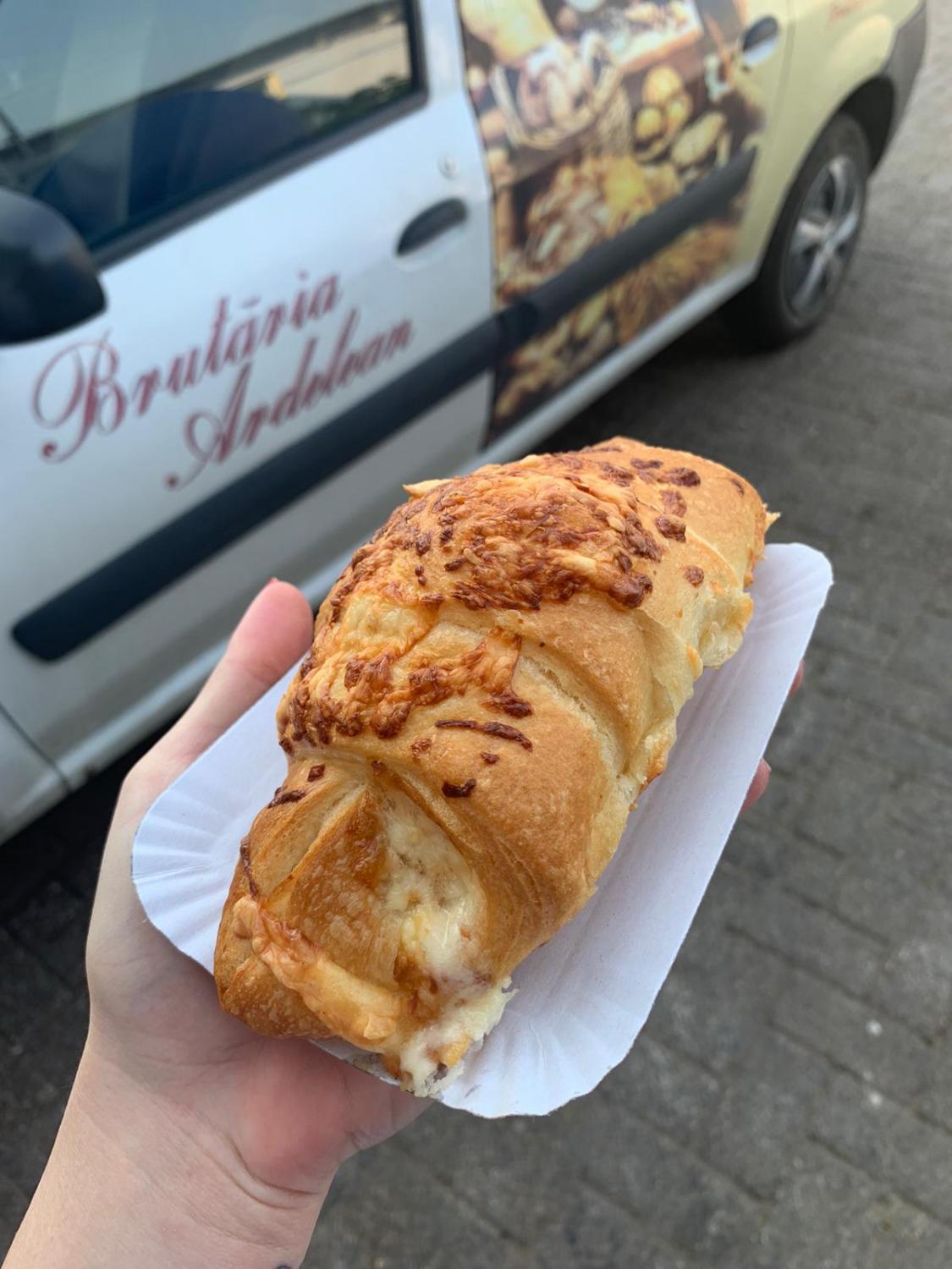 Croissant cu brânză, ținut în mână lângă mașina Brutăriei Ardelean